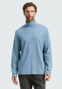 Helles blaues Rollkragensweatshirt aus weichem Stoff, mit langen Ärmeln und entspannter Passform. Kombiniert mit dunkelgrauen Hosen.