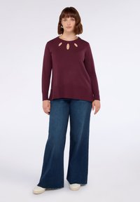 Vinröd långärmad tröja med nyckelhålsutskärningar vid halsen, ihop med vida blå jeans och vita skor. Mjuk textur, avslappnad passform.