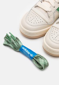 Adidas-tennarit ovat valmistettu beigeistä nahasta, jossa on reikäkuvioita, vihreitä yksityiskohtia ja ruskea kumipohja. Mukana on vihreät ja valkoiset kuvioidut kengännauhat.