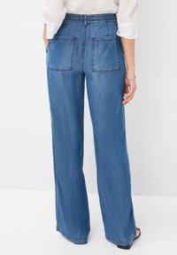 Persona que lleva pantalones de mezclilla azul de pierna ancha con cinturilla elástica y bolsillos de parche, combinados con una blusa blanca de manga larga.