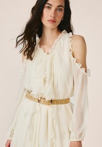 Frau trägt ein cremefarbenes Cold-Shoulder-Kleid mit Rüschen und einem beigen Gürtel mit goldenen Nieten, stehend vor einem schlichten Hintergrund.