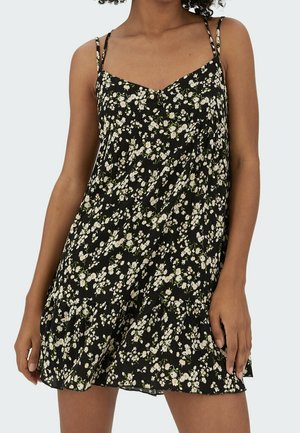 Robe mini noire sans manches avec fines bretelles et motif floral marguerite blanche portée par une personne aux cheveux bouclés, vue du cou jusqu'à mi-cuisse.