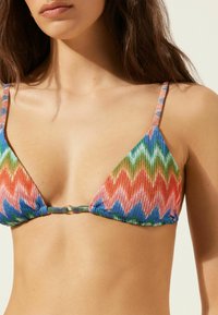 Trojúhelníková bikiny podprsenka vyrobená z texturované látky s barevným zigzag vzorem v červené, modré, zelené a zlatých akcenty. Zlaté kovové detaily vpředu.