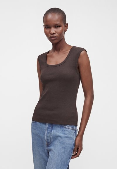 Femme aux cheveux courts portant un haut sans manches marron foncé et un jean bleu clair, debout devant un fond blanc uni.