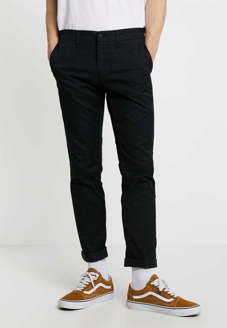 Zwarte slim-fit broek met een gladde textuur en een vijf-pocket ontwerp, gecombineerd met bruine suède sneakers en witte crew-sokken, met opgerolde mouwen zichtbaar.