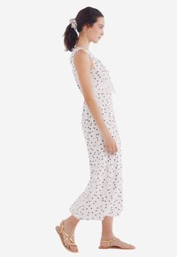 Witte jurk met zwarte stippen, met ruffles en zonder mouwen. Combinatie met bruine platte sandalen en een bijpassende haarband met stippen.