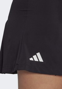 Juoda sportinė sijonas su sluoksniuota struktūra, lygia tekstūra ir baltu Adidas logotipu šalia apvado su trimis įstrižais dryželiais.