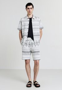 Jeune homme portant une chemise noire, une chemise à boutons à manches courtes à motif blanc et un short assorti, des sandales noires, se tenant avec les mains dans les poches.