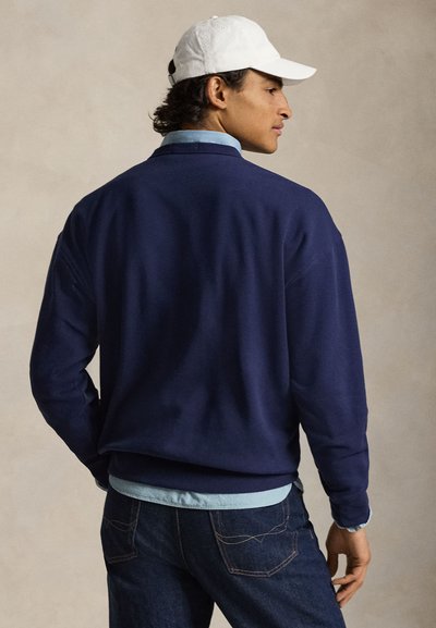 Βαμβακερό φουτερ σε ναυτικό μπλε χρώμα με χαλαρή εφαρμογή, στρογγυλή λαιμόκοψη και ριμπ μανσέτες. Φοριέται πάνω από ανοιχτό μπλε πουκάμισο, σε συνδυασμό με σκουρόχρωμο τζιν και λευκό καπέλο.