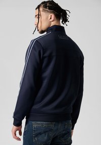 Veste zippée bleu marine en tissu doux avec des rayures blanches contrastantes le long des manches et un détail de logo dans le dos. Assortie à un jean bleu.