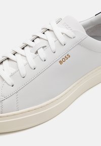 Vita sneakers i slätt läder med platta vita snören, guldbranding på sidan och en beige gummisula. Minimalistisk design.