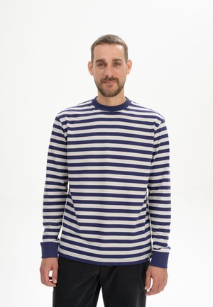 Man met kort haar en baard, gekleed in een blauw-wit gestreept shirt met lange mouwen en een zwarte broek, staand tegen een effen witte achtergrond.