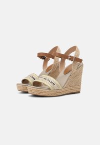 Beige kilehæle sandaler med en flettet jute sål, brun ankelrem, og stofbånd med "TOMMY HILFIGER" tekst i sort og gule accenter.