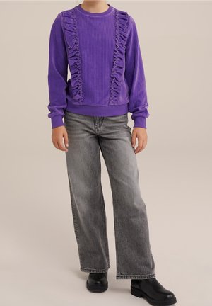 Kind draagt een paarse trui met ruches en lange mouwen, grijze wijde jeans en zwarte enkellaarzen, staand tegen een effen achtergrond.