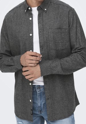 Homme portant une chemise gris foncé à boutons par-dessus un t-shirt blanc et un jean bleu, en train de boutonner le bouton du milieu de la chemise.