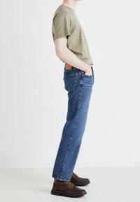 Olivgrön bomull t-shirt, blå denimjeans, bruna snörkängor. Jeansen har en rak passform med en läderlapp på midjan.