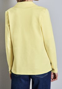 Lange mouwen shirt van zacht gele katoen, met een kraag en afgeronde zoom, gecombineerd met blauwe spijkerbroek. De stof heeft een gladde textuur.