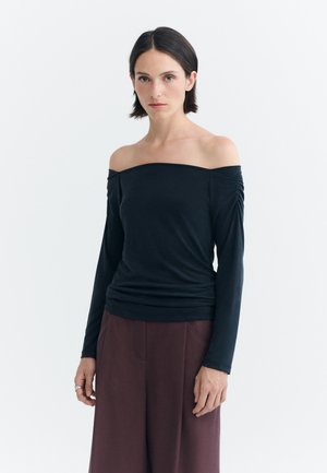 Jeune femme aux cheveux courts et foncés portant un top noir à manches longues et épaules dénudées ainsi qu'un pantalon large marron foncé, debout sur un fond blanc.