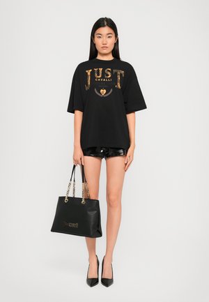 T-shirt noir surdimensionné avec imprimé doré "JUST CAVALLI", associé à un short noir brillant et un sac à main noir avec des anses en chaîne dorée.