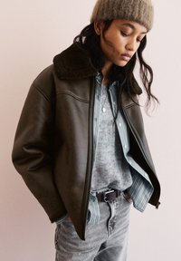 Vrouw met een bruine schapenvachtjas, gestreept shirt, grijze trui, grijze spijkerbroek en een beige gebreide muts, kijkend naar beneden met handen in de zakken.