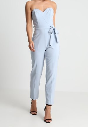 Sukienka bez ramiączek w jasnoniebieskim kolorze z dekoltem w kształcie serca i paskiem w talii, zestawiona z czarnymi sandałami na wysokim obcasie, na stojącym modelu.