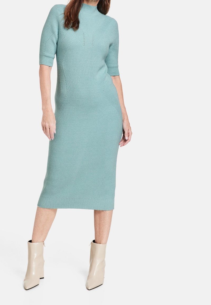 Femme portant une robe midi en maille côtelée bleu clair avec des manches mi-longues jusqu'aux coudes et des bottines beige à talons sur un fond blanc.