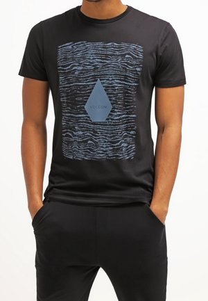 Mann trägt ein schwarzes T-Shirt mit welligem blauem Muster und einer blauen Diamantform mit dem Text "VOLCOM" in der Mitte.