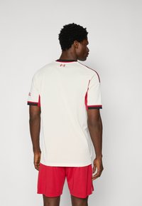 Valge spordijakak, millel on punased aktsendid ja must äär, lühikesed varrukad, ümmargune kaelus. Seljal on väike logo detail.