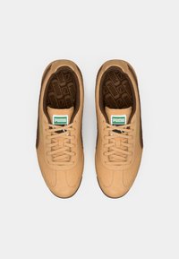 Καφέ σουέτ αθλητικά παπούτσια με μπεζ λεπτομέρειες, διαθέτοντας υφή στη γλώσσα, μαύρα κορδόνια και πράσινη ετικέτα λογότυπου στο μπροστινό μέρος.