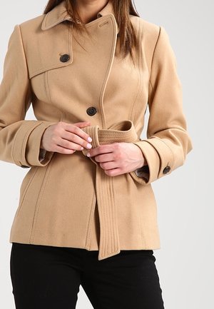 Kvinde iført beige dobbeltradet frakke med sorte knapper, justerer matchende bælte, kombineret med sorte bukser mod en lys grå baggrund.