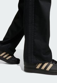 Čierne športové nohavice s tromi pruhmi po stranách, v kombinácii s čiernymi topánkami s textúrovaným dizajnom a tromi béžovými pruhmi.