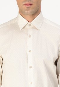 Cămașă button-up bej deschis, cu un model geometric subtil, guler clasic și textură catifelată, având cinci butoane în față.