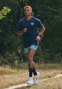Zila skriešanas krekls, gaiši zili šorti, melnas sporta zeķes un baltas ar krāsainiem akcentiem skriešanas kurpes. Brīvā dabā uz zemes ceļa.