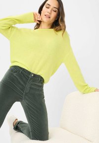 Heller limonengrüner, übergroßer Pullover kombiniert mit dunkelgrünen Cordhosen. Das Model lehnt an einem strukturierten cremefarbenen Sitz.