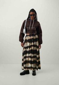 Vrouw in een donkerbruine cropped hoodie, zwarte en beige gemêleerde lange rok, zwarte laarzen, zonnebril en een zilveren schoudertas, staande.
