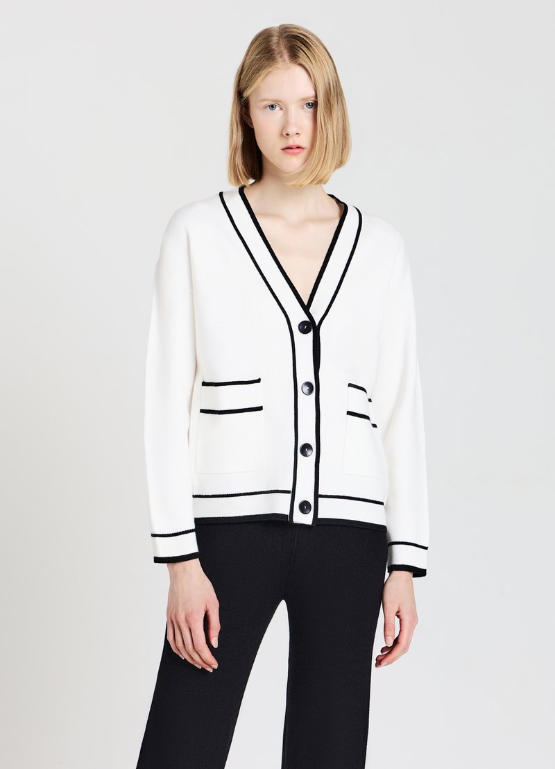 Cardigan bianco con rifiniture nere, scollo a V e tasche frontali. I bottoni anteriori ne migliorano la funzionalità. Texture morbida e vestibilità rilassata.