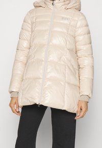 Beżowa pikowana kurtka puchowa z kapturem, zamkiem błyskawicznym z przodu, bocznymi kieszeniami i ściągaczami na mankietach, noszona z czarnymi spodniami.