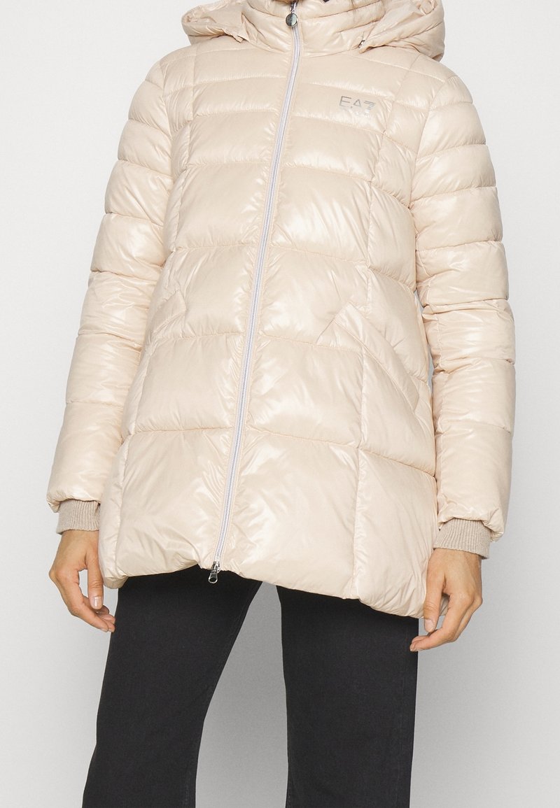 Beżowa pikowana kurtka puchowa z kapturem, zamkiem błyskawicznym z przodu, bocznymi kieszeniami i ściągaczami na mankietach, noszona z czarnymi spodniami.