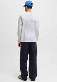 Man in een blauwe pet, wit langemouwen shirt, zwarte wijde broek en beige schoenen, staande met de rug naar de camera op een witte achtergrond.