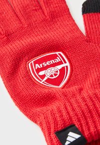 Røde strikkede handsker med et fremtrædende hvidt Arsenal-logo på en skjoldbaggrund. Sort kant ved håndleddet med Adidas-branding.