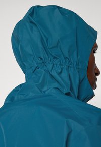 Τζάκετ σε χρώμα τιρκουάζ με κουκούλα, φτιαγμένο από ελαφρύ, υφασμάτινο υλικό. Διαθέτει συγκεντρωμένη ελαστική ταινία στην κουκούλα για τέλεια εφαρμογή. Ορατή φερμουάρ στο πλάι.