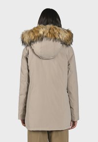 Béžová parka bunda s kapucňou so kožušinovým lemom. Má dlhé rukávy a rovný strih. Kožušina je v kombinácii hnedých a pieskových farieb.