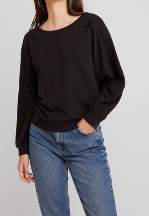 Mujer con sudadera negra de manga larga con bordado floral sutil en las mangas y jeans de mezclilla azul, con la mano en el bolsillo, fondo neutro.