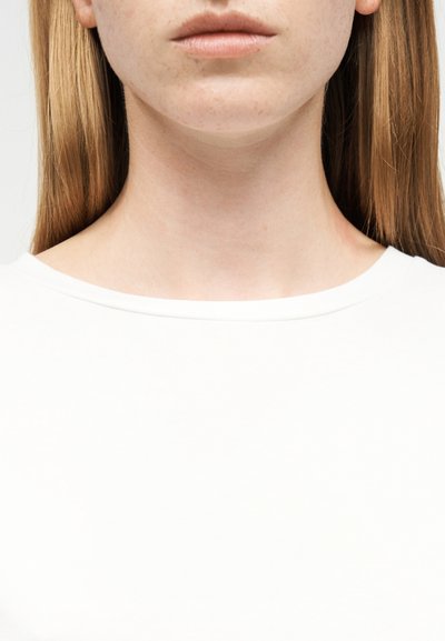 Κοντινό πλάνο στο πηγούνι, τον λαιμό και την περιοχή του ώμου ενός ατόμου με μακριά, ίσια μαλλιά που φοράει λευκό μπλουζάκι με στρογγυλή λαιμόκοψη, σε απλό φόντο.