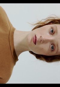 Mladić svijetle puti i crvene kose obučen u smeđu košulju, gleda izravno u kameru na jednobojnoj bijeloj pozadini.