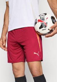 Rote Sportshorts mit einem orangen Seitenstreifen und einem Puma-Logo. Der Stoff wirkt leicht und atmungsaktiv. Ein Fußball wird in der Hand gehalten.