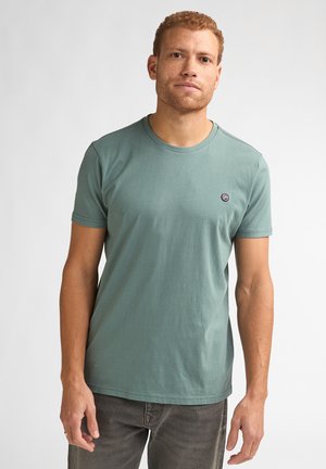 Man with short curly hair wearing a fitted sage green short-sleeve t-shirt and dark pants against a plain light background.