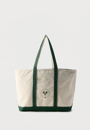 Sac cabas en toile beige avec des poignées vert foncé, une base et un logo de raquette de tennis sur la poche avant, posé devant un fond uni.