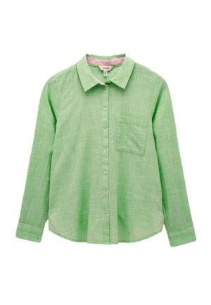 Brazos cruzados con una camisa de manga larga pequeña a cuadros verde claro con botones blancos y texto bordado parcialmente visible en el puño.