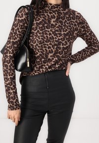 Langärmliges Oberteil mit Leopardenmuster in Braun und Beige, kombiniert mit schwarzen, enganliegenden Hosen. Die Textur ist glatt mit einem leichten Glanz. Schwarze Handtasche mit Reißverschlussdetails.